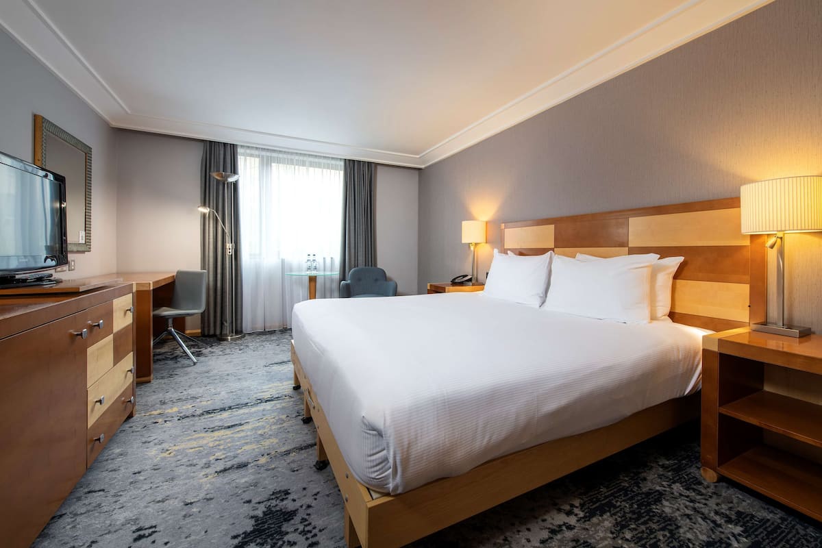 A hotel room with a large bed, a desk, a chair, a TV, and a lamp.