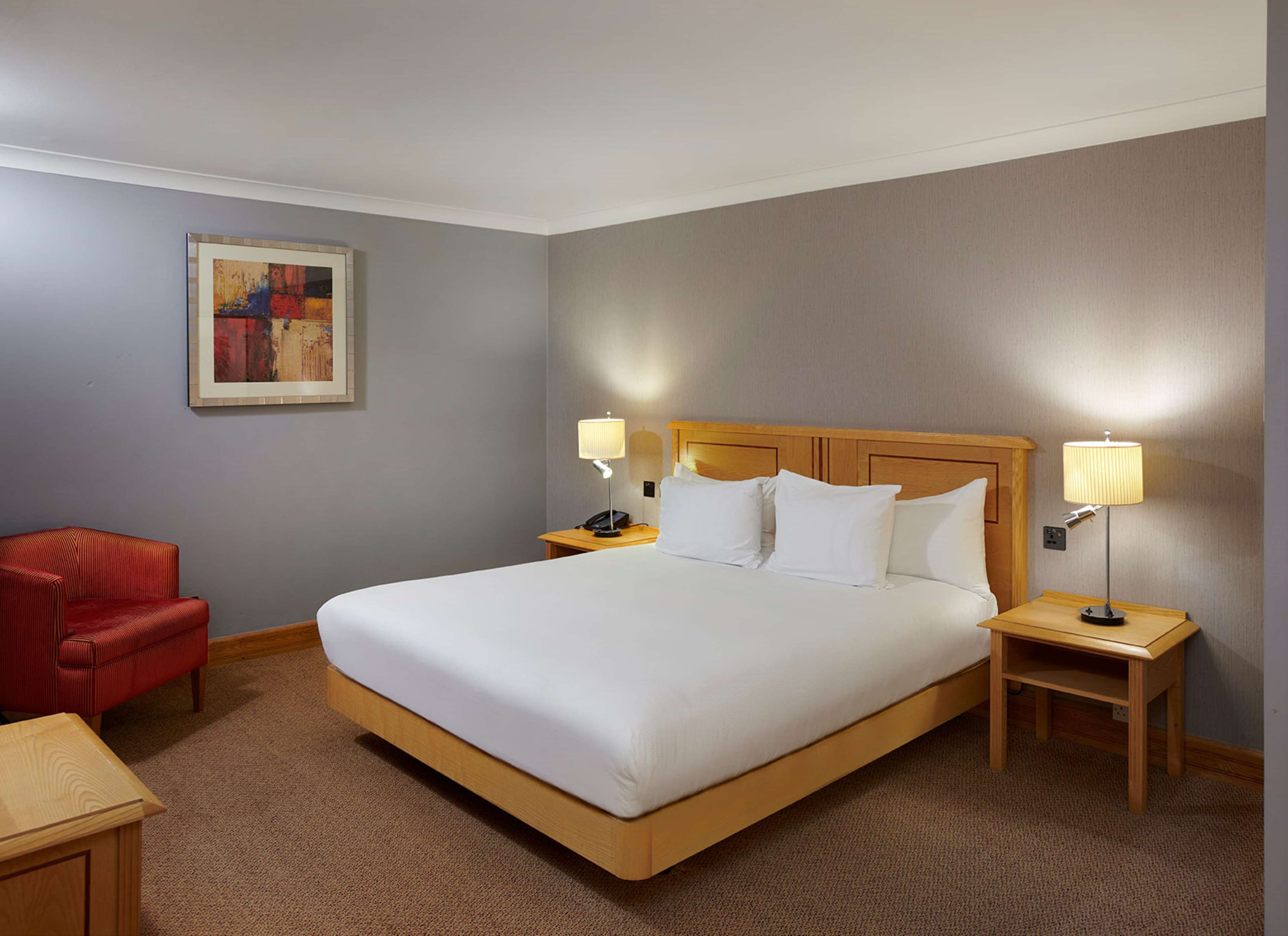 A hotel room with a large bed, two bedside tables with lamps, a red chair, and a painting on the wall.
