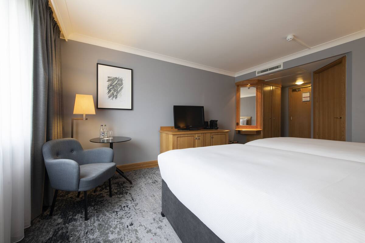 A hotel room with a large bed, a chair, a small table with a lamp, a television, and a wardrobe.