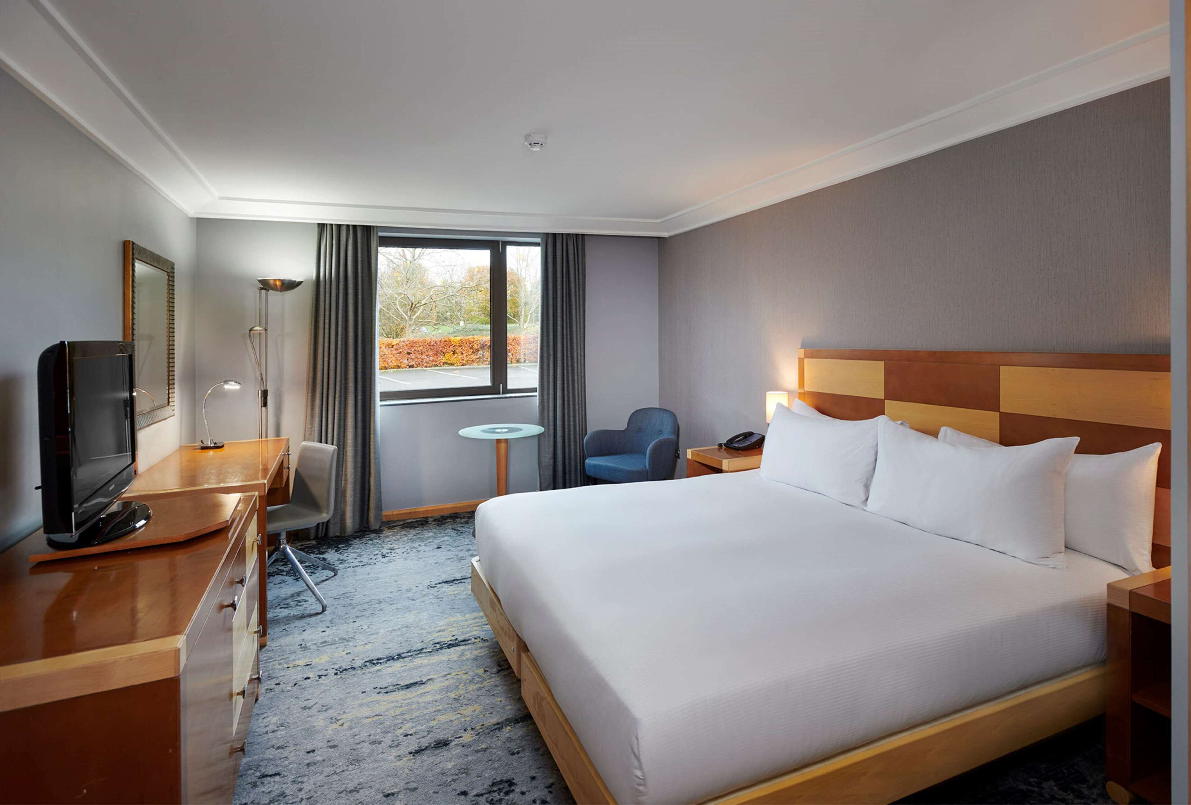 A hotel room with a large bed, a desk with a television, a chair, a small table, and a window with a view of trees.