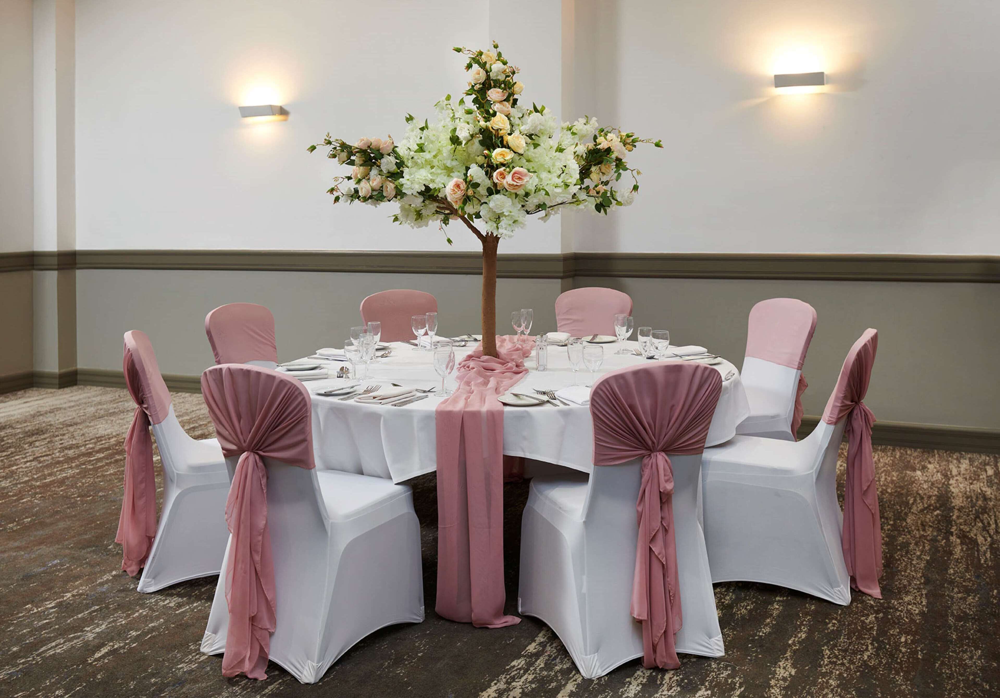 A round table with white tablecloths and pink chair covers, surrounded by white chairs.