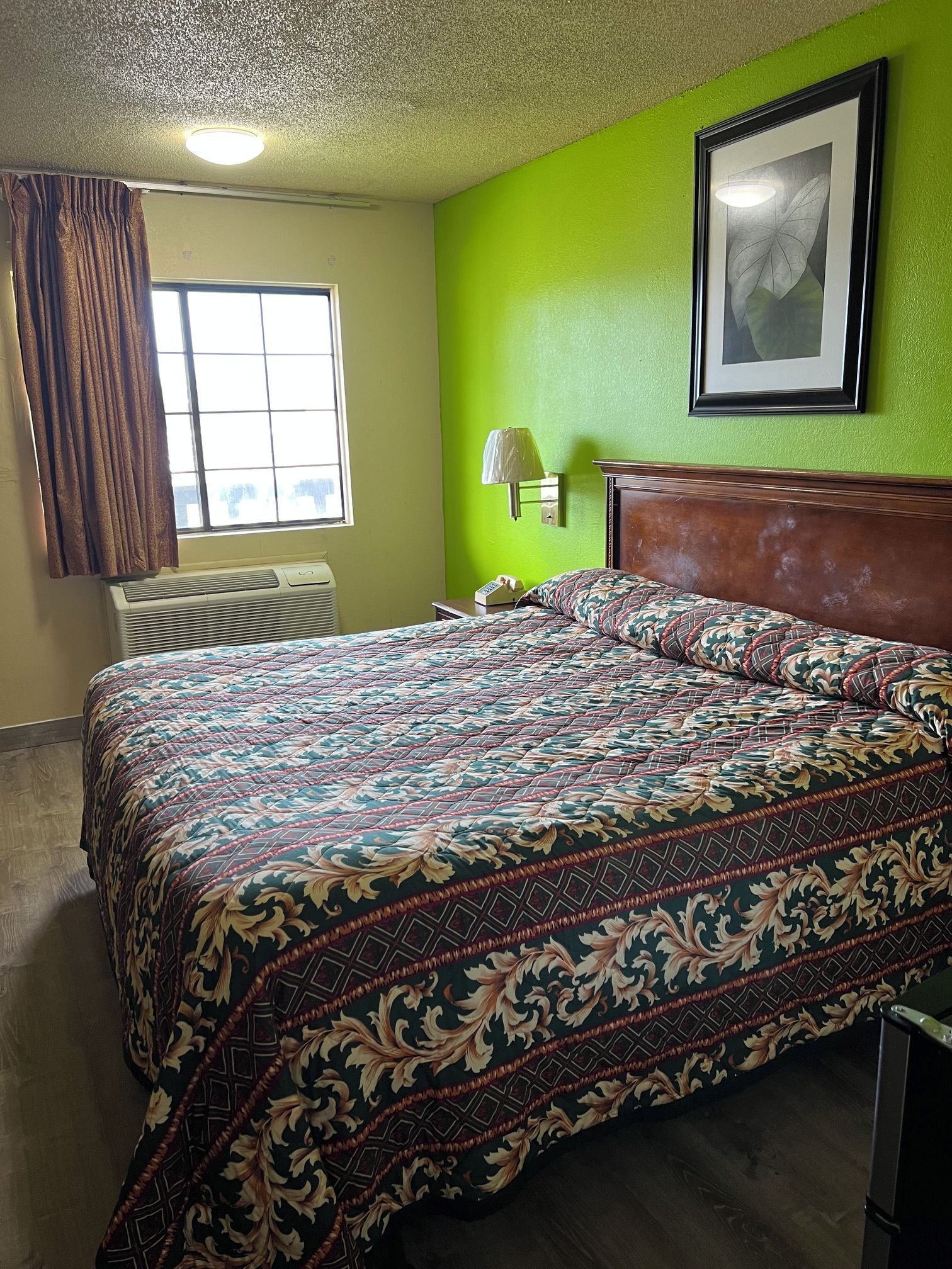 A hotel room with a bed, a nightstand, a lamp, a framed picture on the wall, and a window with curtains.