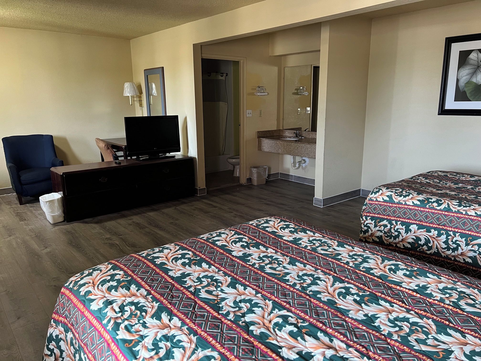 A hotel room with a bed, a desk with a television, a chair, a bathroom with a sink and mirror, and a framed picture on the wall.