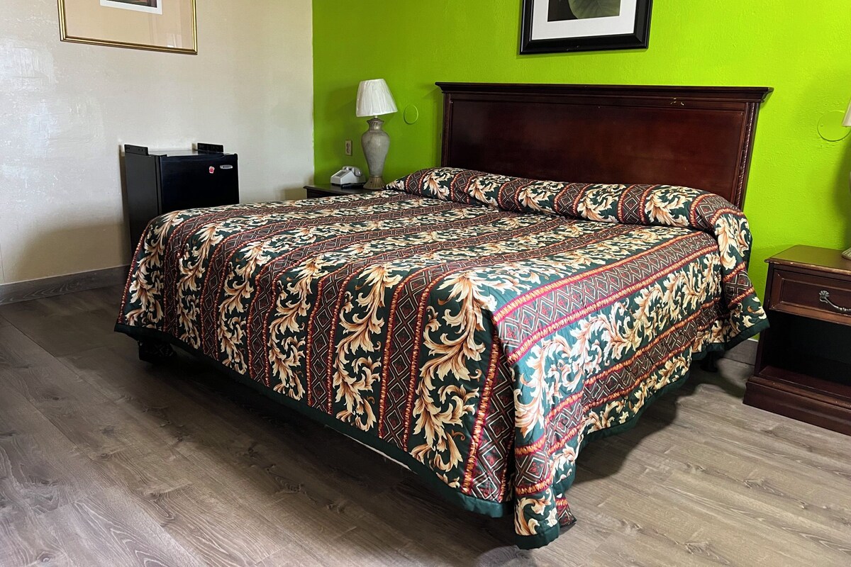 A hotel room with a large bed, a wooden headboard, a nightstand, a lamp, and a framed picture on the wall.