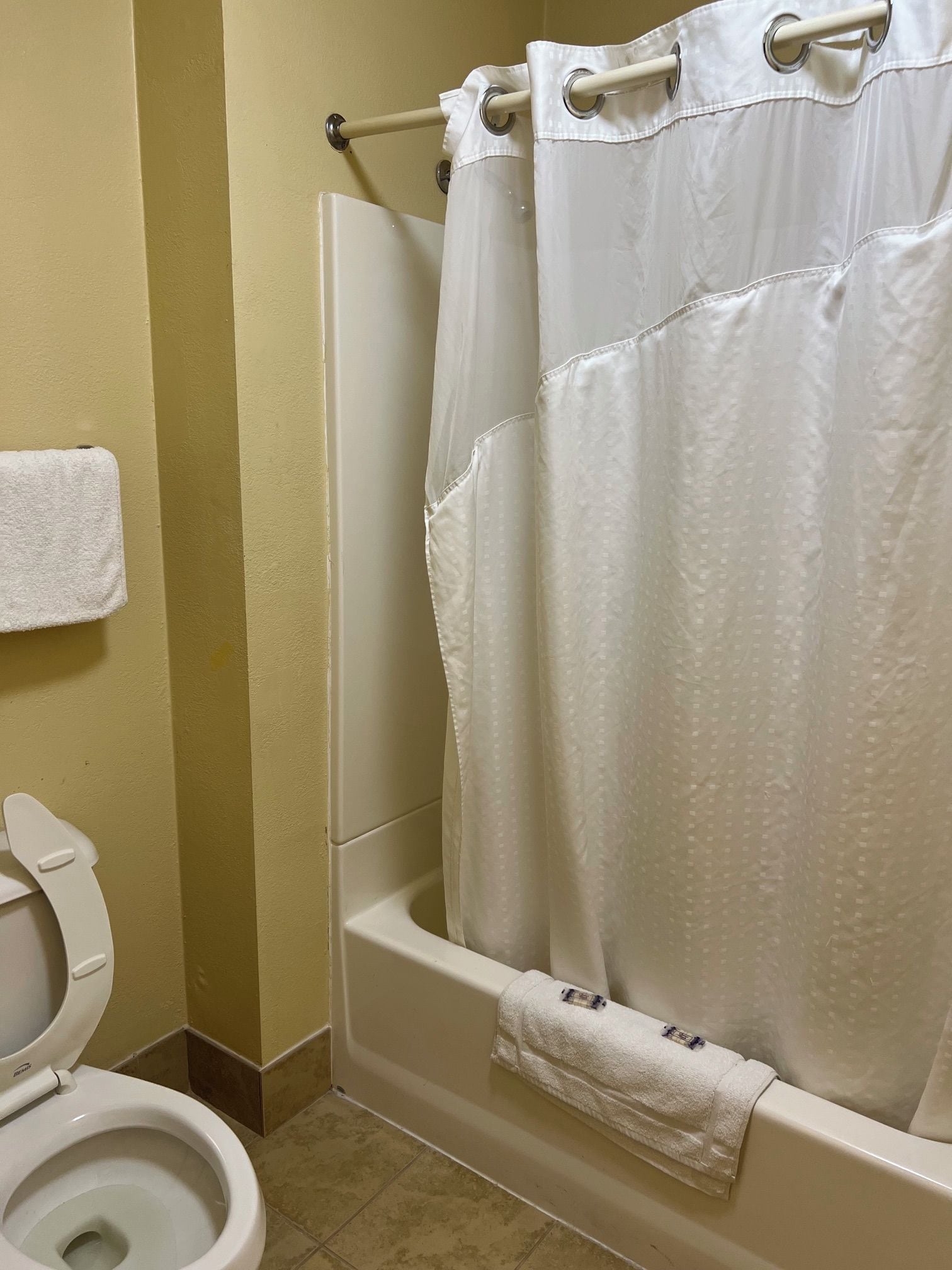 Un baño con inodoro blanco, bañera con cortina de ducha blanca y una toalla en el suelo.