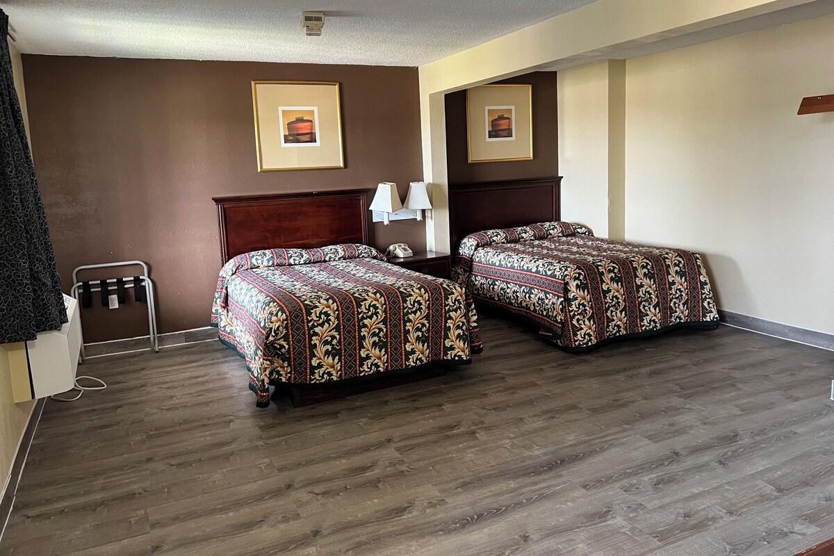A hotel room with two beds, a wooden headboard, a bedside table with a lamp, and a wall-mounted air conditioning unit.
