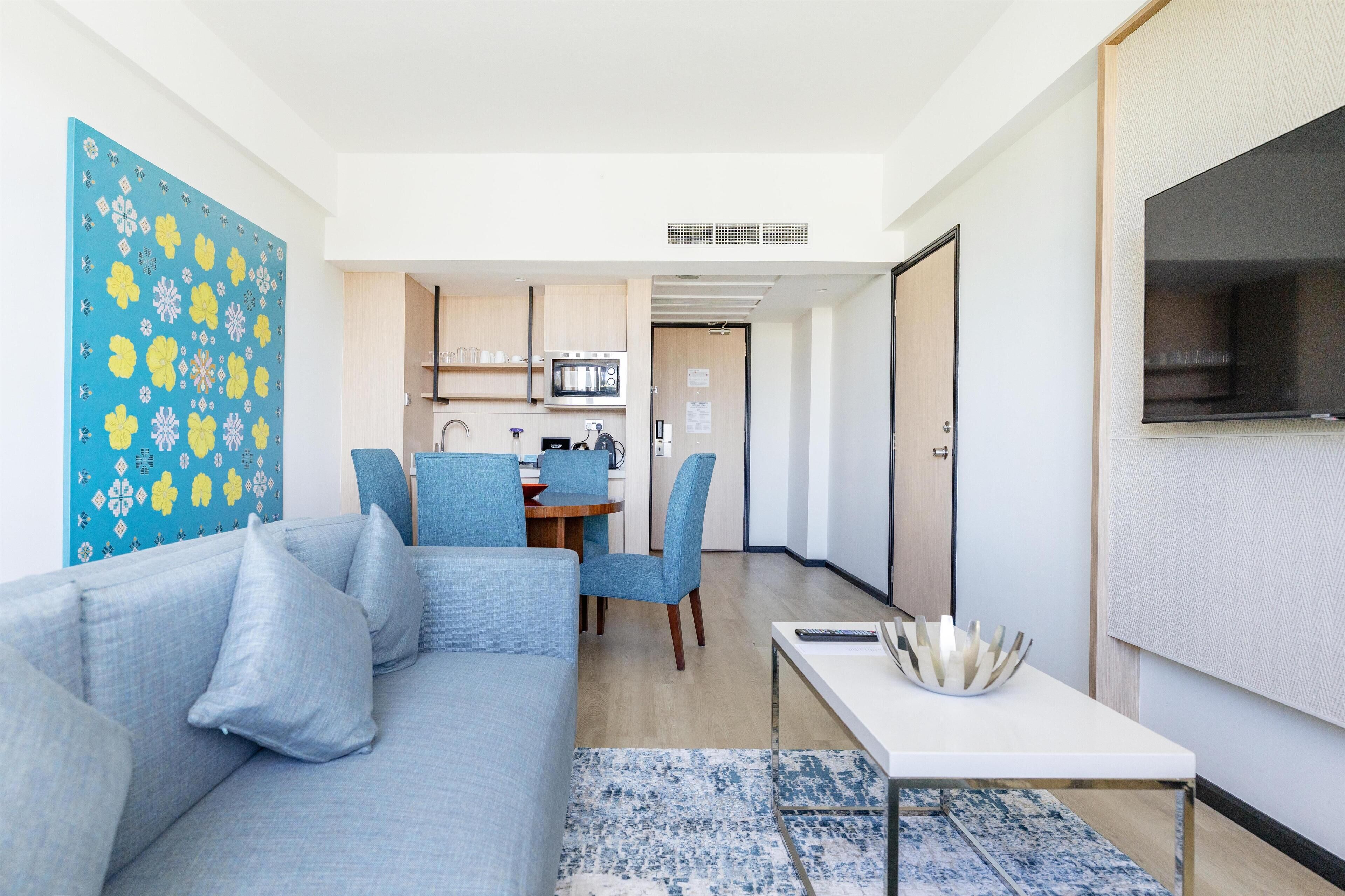 A modern living room with a sofa, a dining area with chairs, a kitchen with appliances, and a TV mounted on the wall.