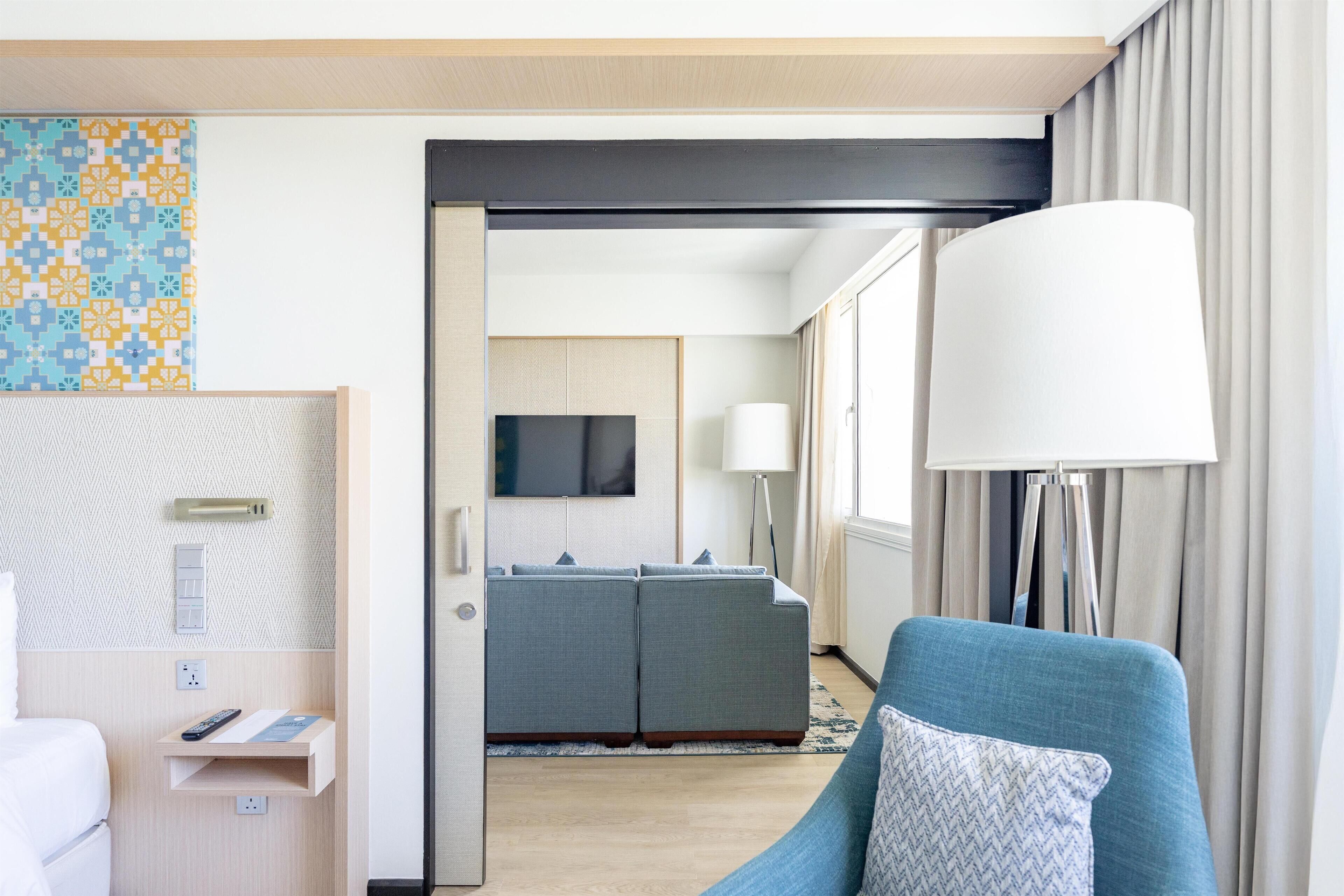 A hotel room with a blue sofa, a white armchair, a TV, a bedside table with a lamp, and a patterned wall.