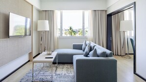 Habitación de hotel moderna con un sofá gris, una mesita pequeña, un televisor montado en una pared texturizada y vistas a palmeras a través de grandes ventanales.