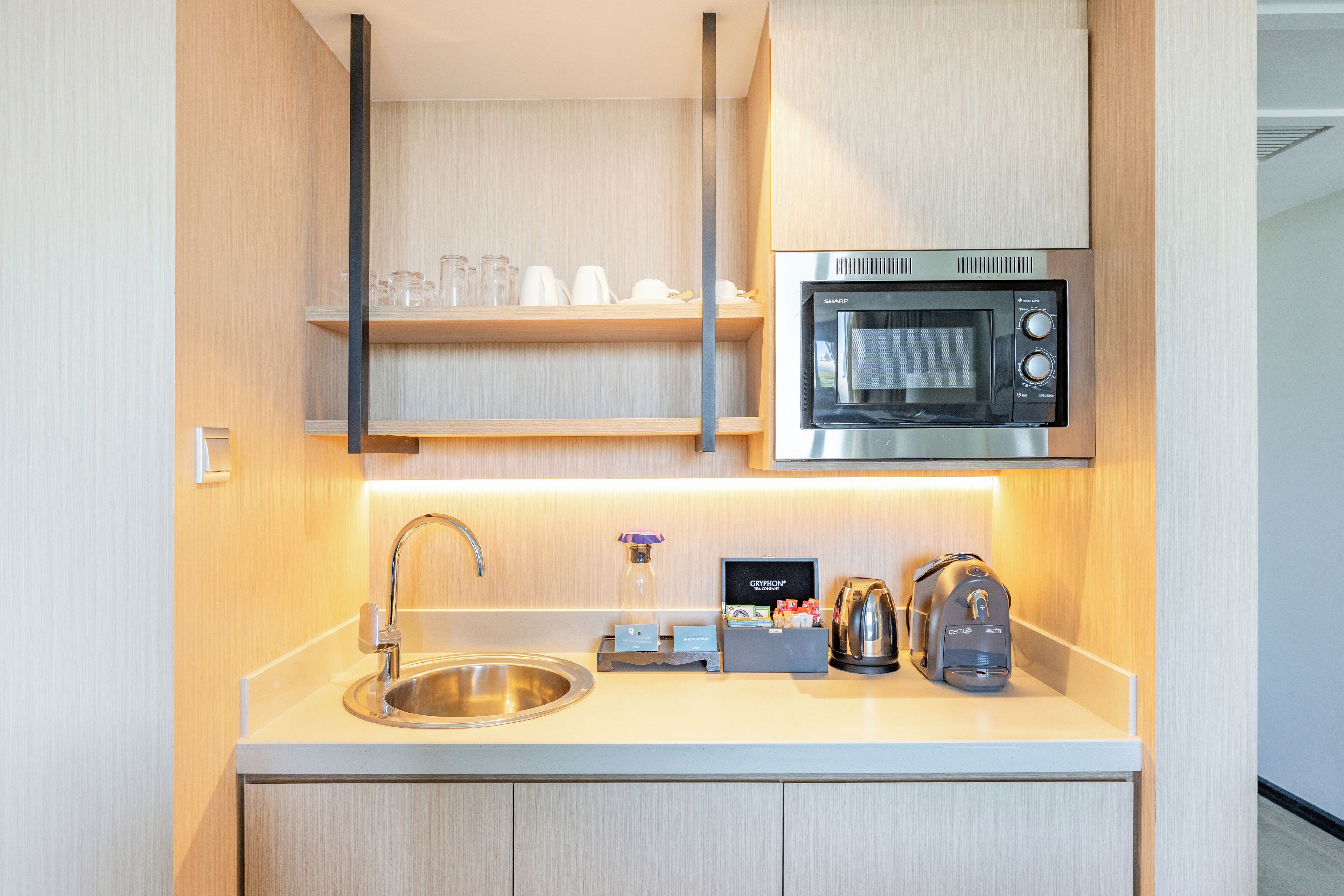 A modern kitchen with a stainless steel sink, a microwave, a kettle, and a shelf with glassware.