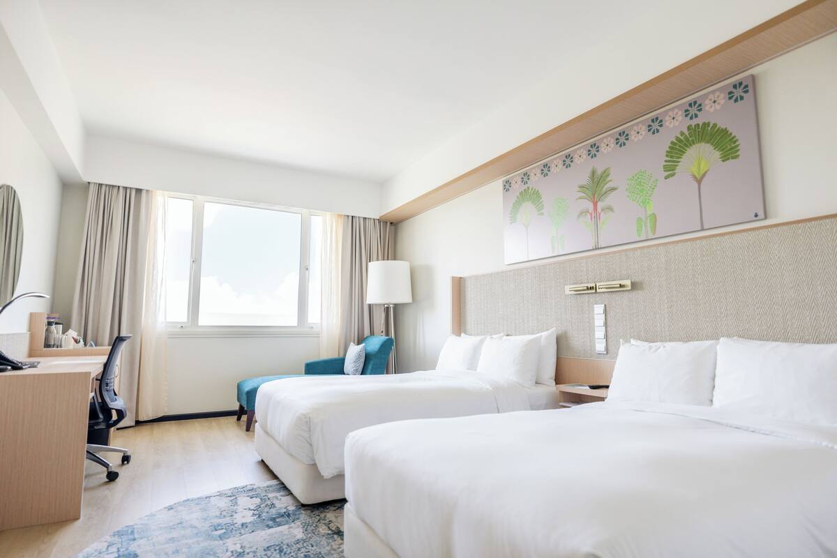 Une chambre d’hôtel avec deux lits, un bureau, une chaise, une lampe et une fenêtre avec des rideaux.