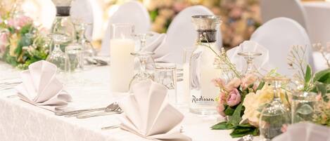 Mesa dispuesta para un evento formal, con manteles blancos, cristalería y centros de mesa florales.