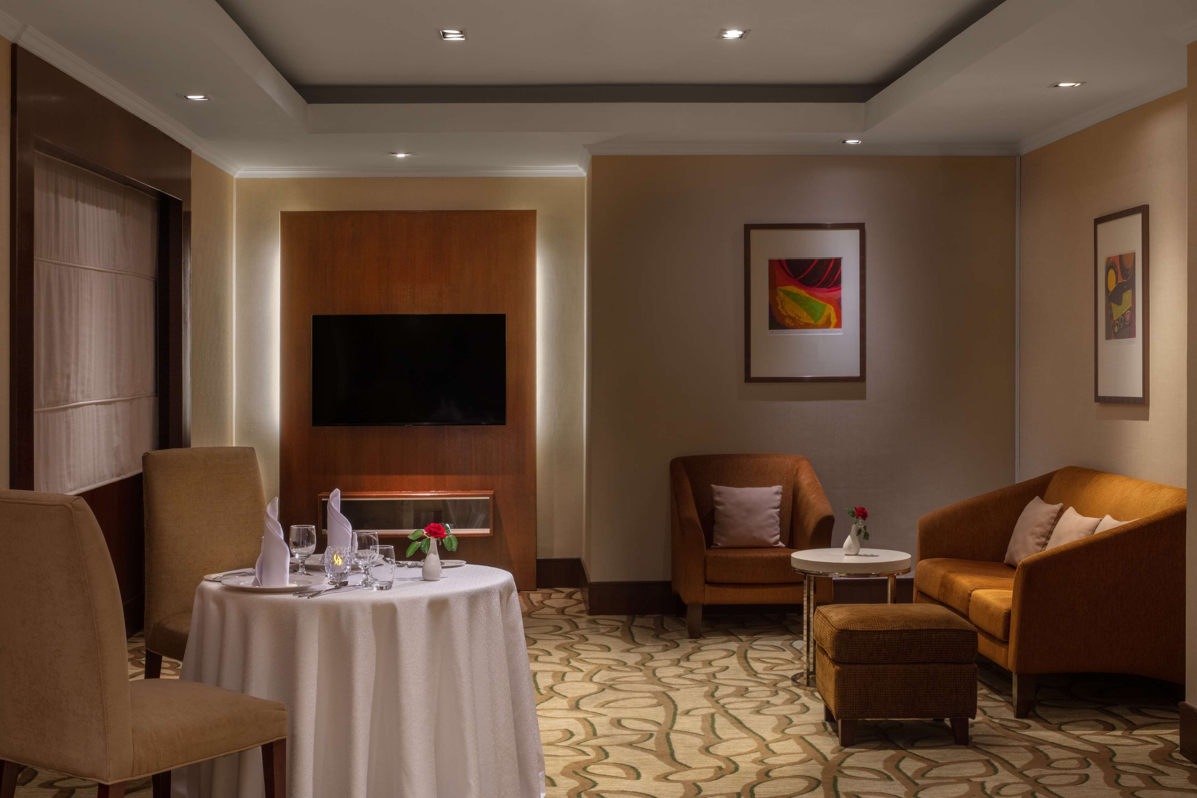 A hotel room with a dining table set for two, a sofa, a small armchair, and a television mounted on the wall.