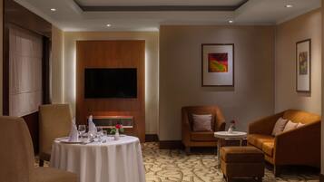 A hotel room with a dining table set for two, a sofa, a small armchair, and a television mounted on the wall.