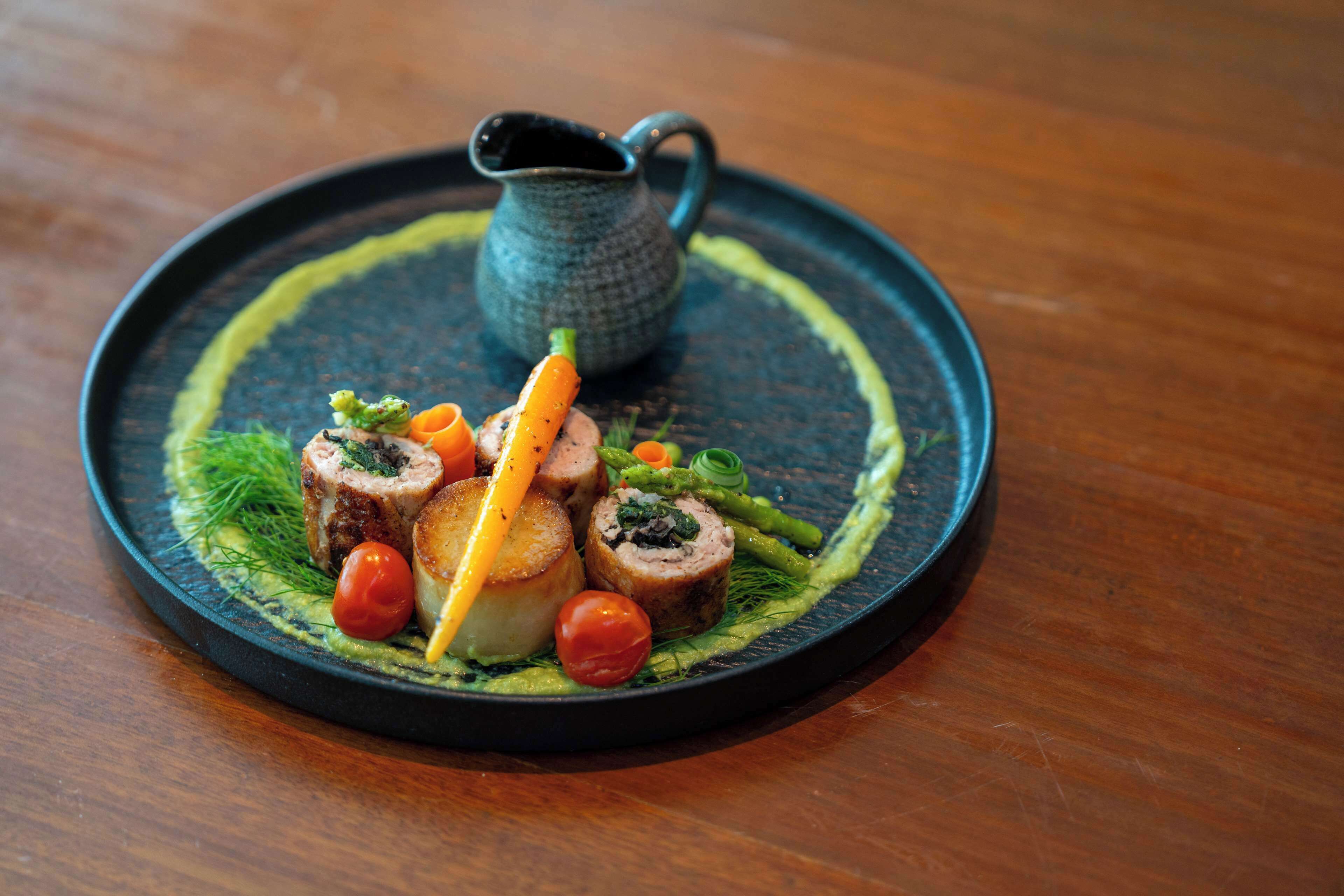 A round black plate with a dish of food, including rolled meat, vegetables, and a small pitcher.