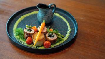 A round black plate with a dish of food, including rolled meat, vegetables, and a small pitcher.