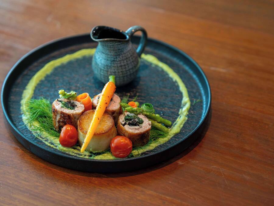 A round black plate with a dish of food, including rolled meat, vegetables, and a small pitcher.