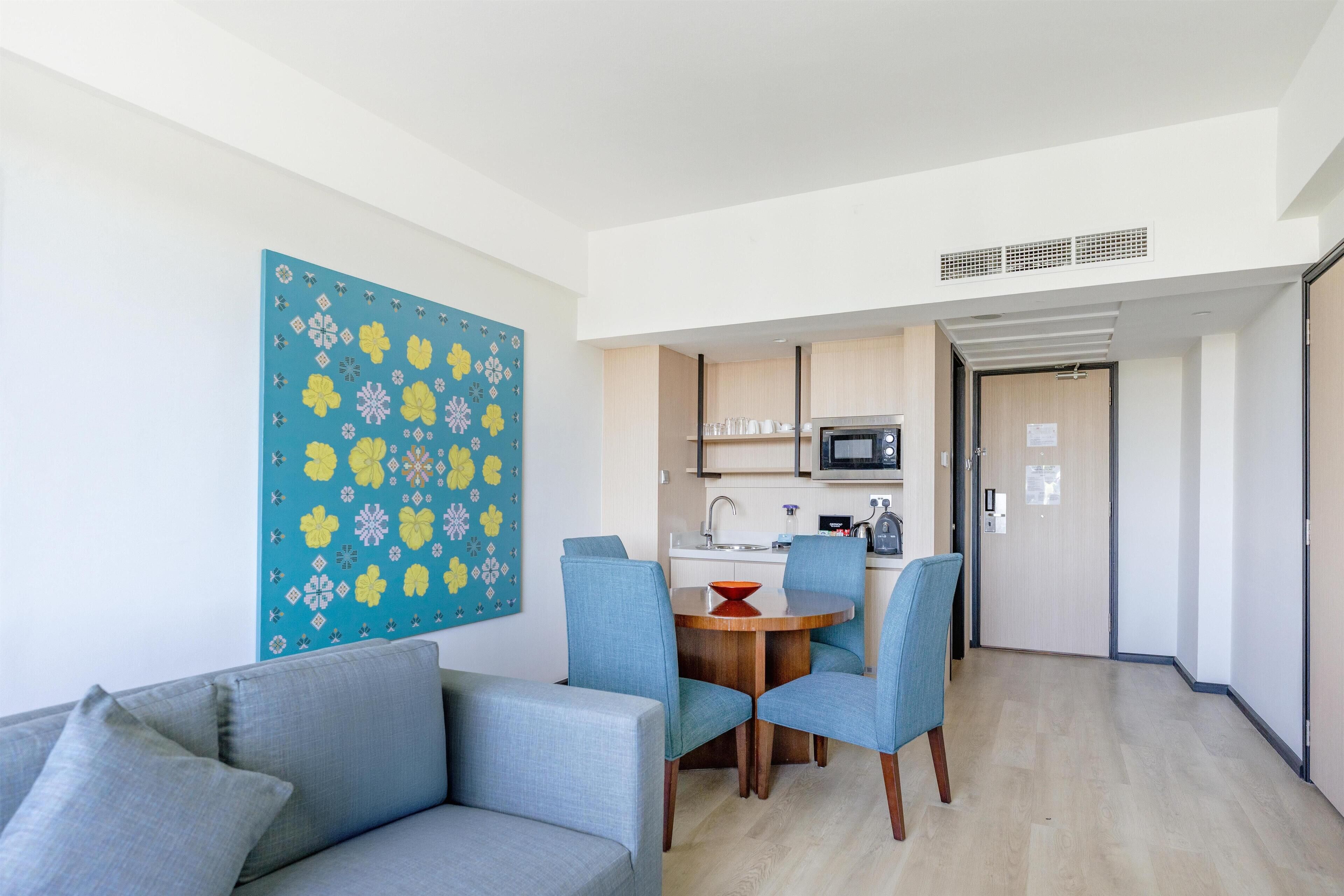 Una sala moderna con sofá y sillones, una mesa de comedor con sillas, una zona de cocina con microondas y una puerta que da a otra habitación.