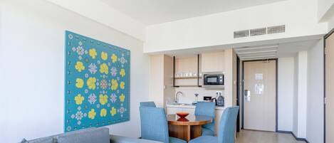 Una sala moderna con sofá y sillones, una mesa de comedor con sillas, una zona de cocina con microondas y una puerta que da a otra habitación.