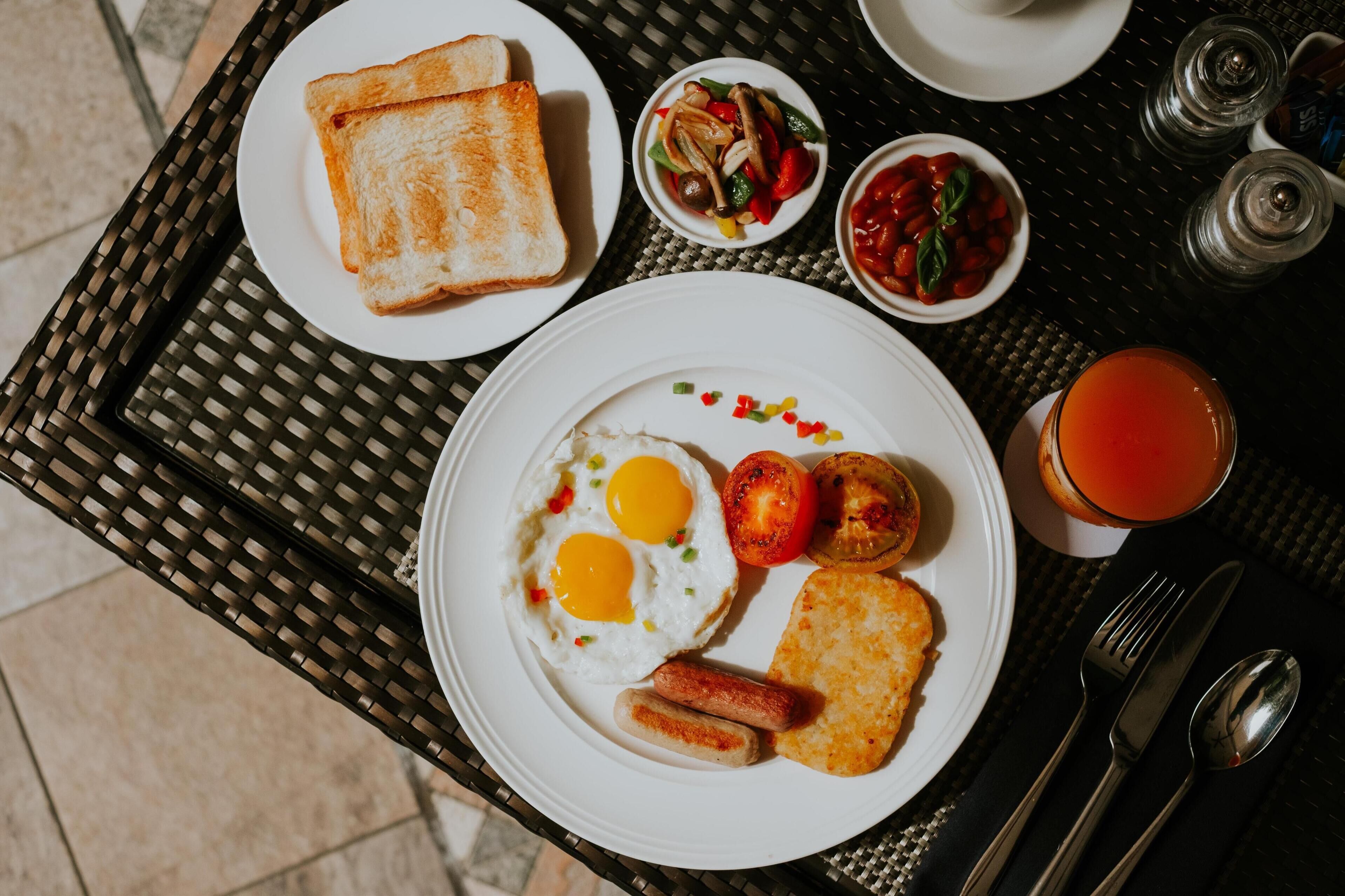Una mesa de desayuno con tostadas, huevos, salchichas, tomates y un vaso de jugo de naranja.
