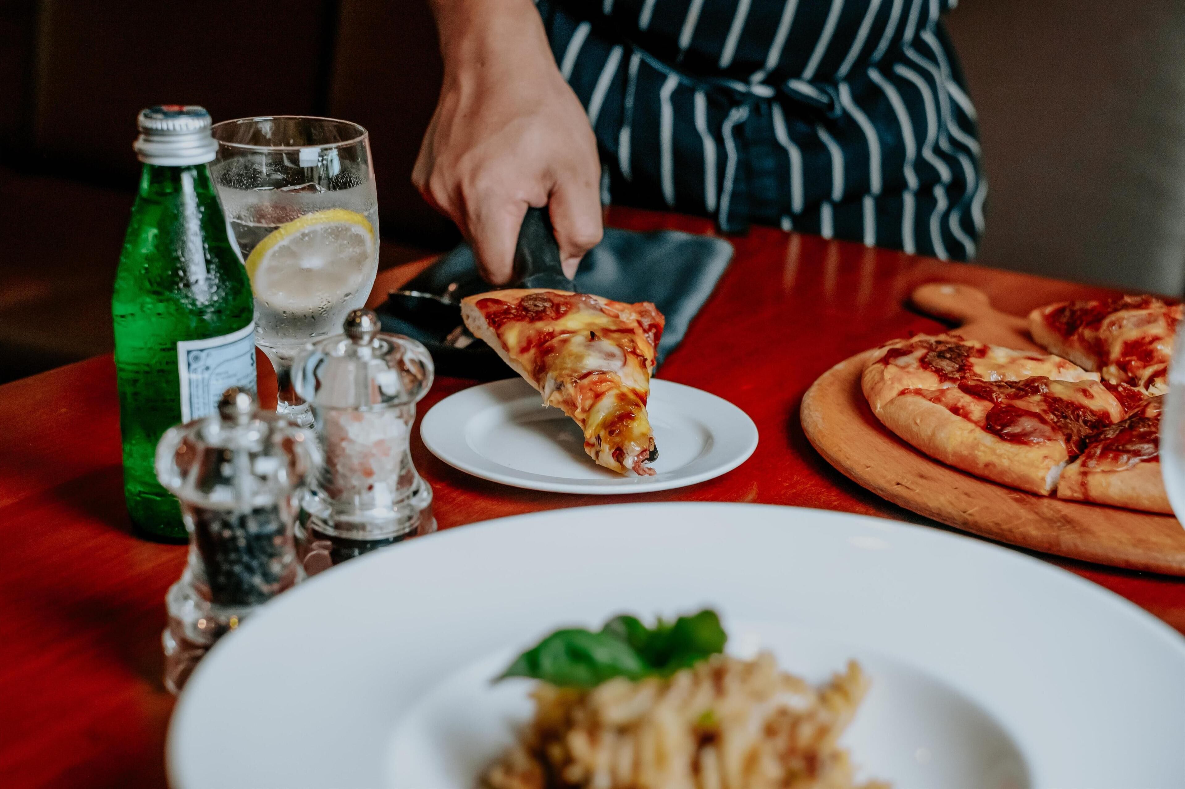 Een tafel met een stuk pizza, een hele pizza, een blikje frisdrank, een glas water en een bord friet.