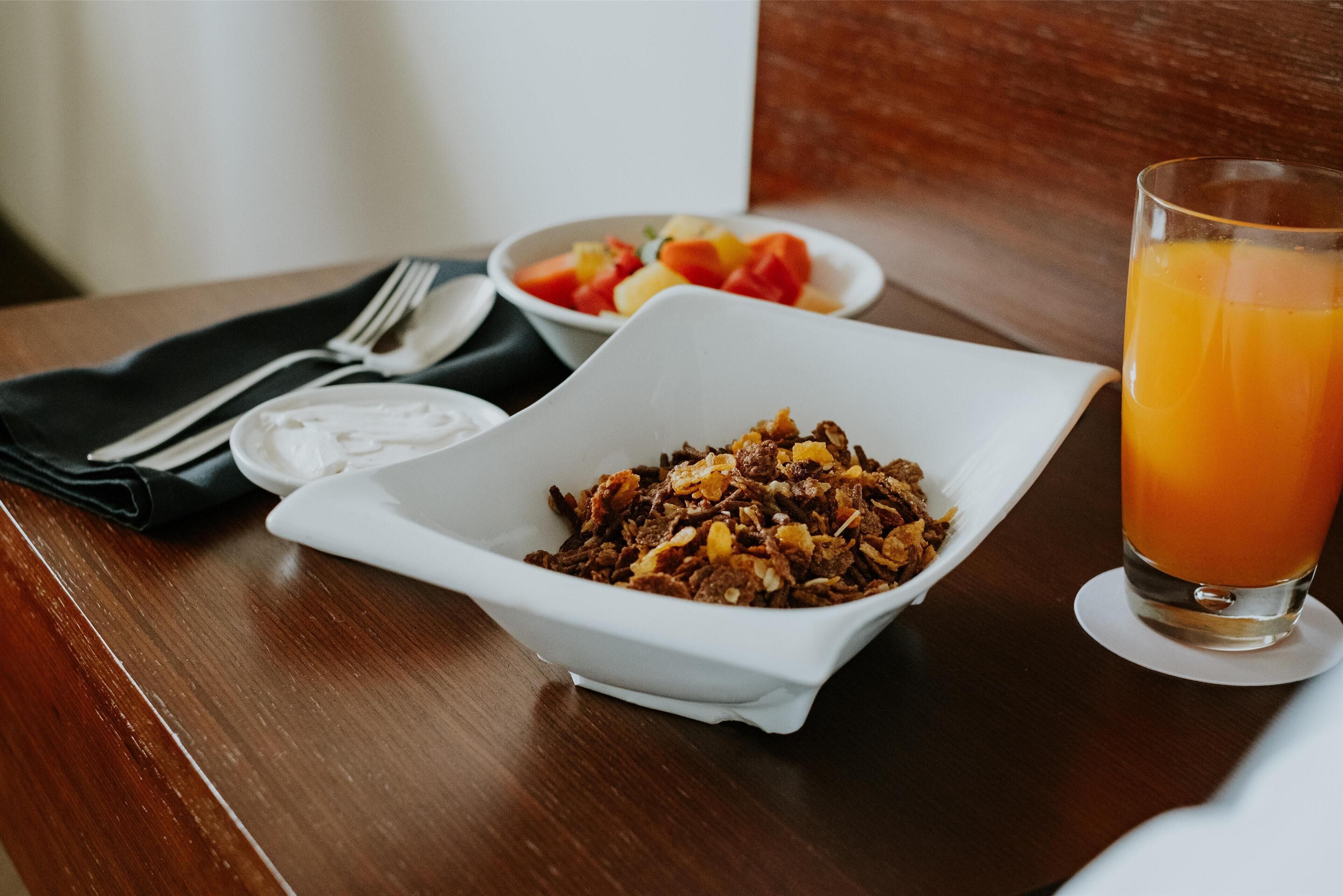 A breakfast setting with a white bowl of cereal, a glass of orange juice, and a plate of fruit on a wooden table.