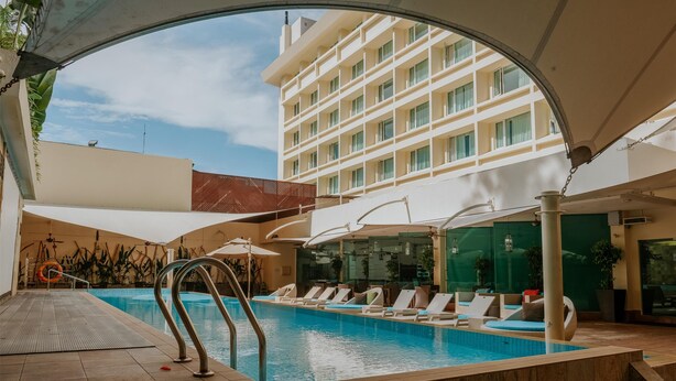 Una piscina con sillas de descanso y sombrillas, rodeada por un edificio de varios pisos y grandes ventanales.