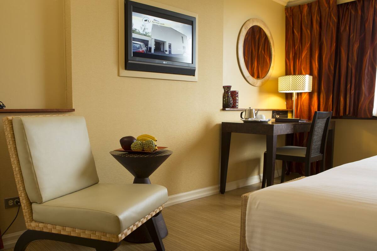 A hotel room with a bed, a chair, a small table with fruit, a lamp, and a television on the wall.