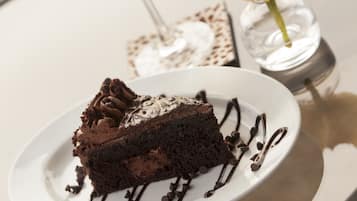 Une part de gâteau au chocolat sur une assiette blanche, nappée d’un filet de chocolat, un verre de vin blanc et une fleur jaune dans un vase en verre, le tout posé sur une table ronde.