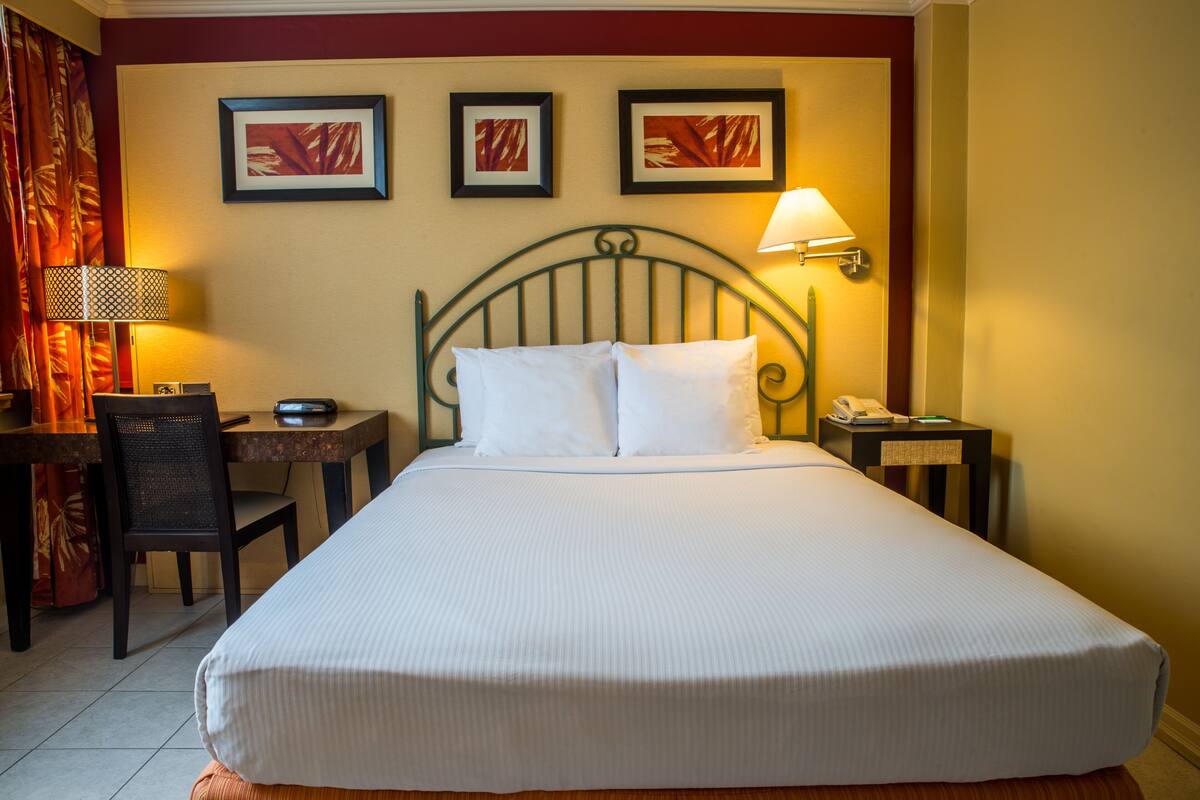 A hotel room with a large bed, a desk with a chair, a lamp, and two framed pictures on the wall.
