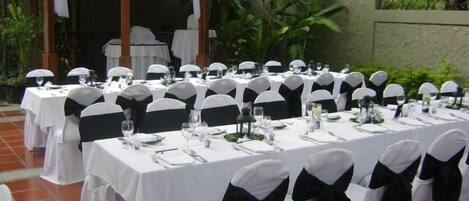 An outdoor dining area with white tables and chairs, each with black sashes, set against a backdrop of greenery and a tiled structure.