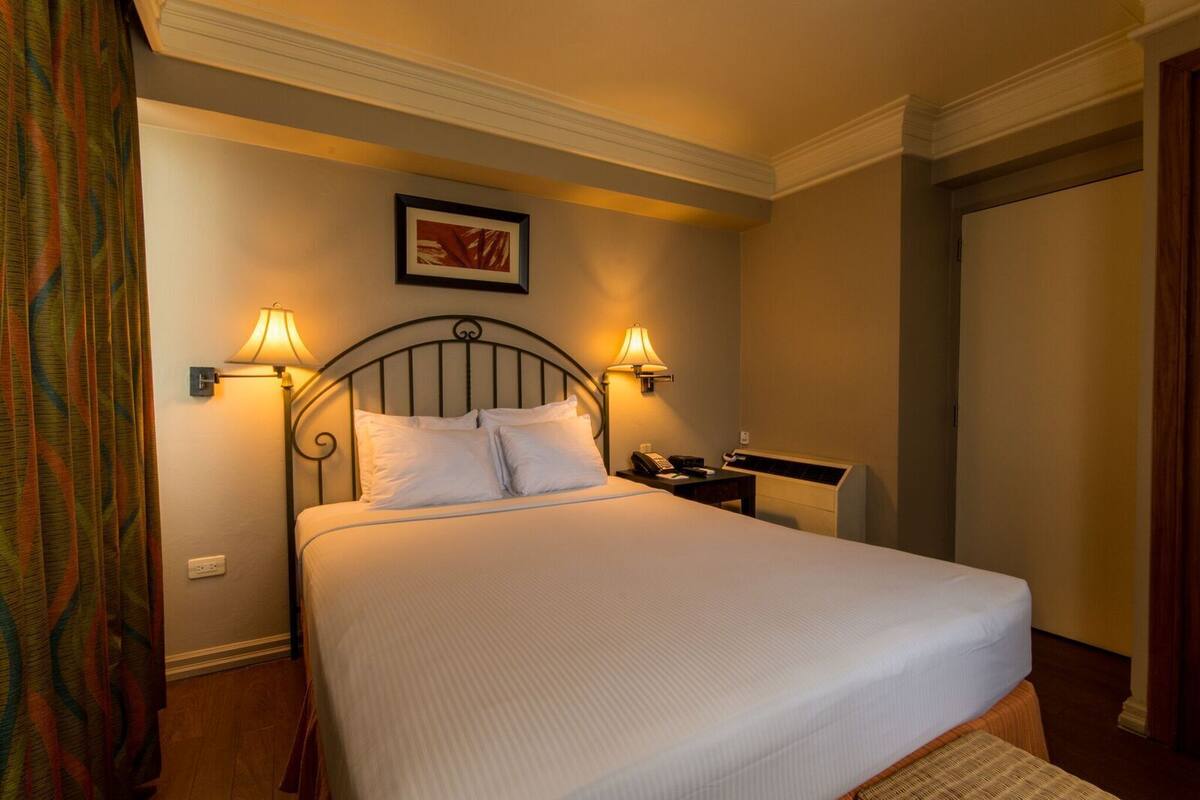 A neatly made bed with white linens, two bedside lamps, a phone on the nightstand, and a framed picture on the wall.
