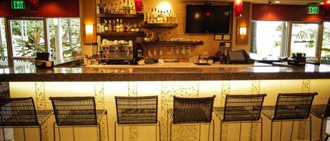 Zona de bar con mostrador, taburetes, estantes con botellas, un televisor y letreros de salida.