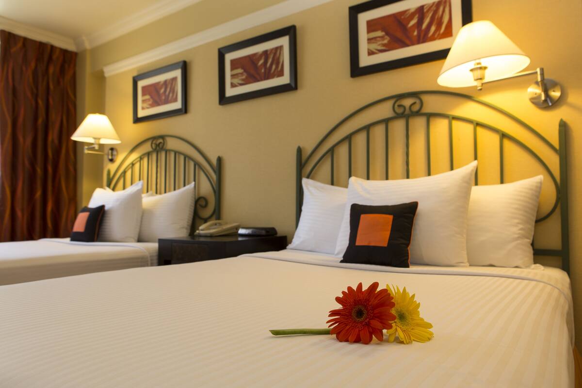 A hotel room with two beds, each with white linens and decorative pillows. There are wall-mounted lamps and framed artwork above the beds.