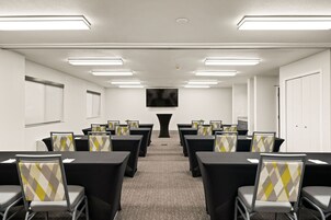 A conference room with rows of tables and chairs, a flat-screen TV mounted on the wall, and a door on the right.