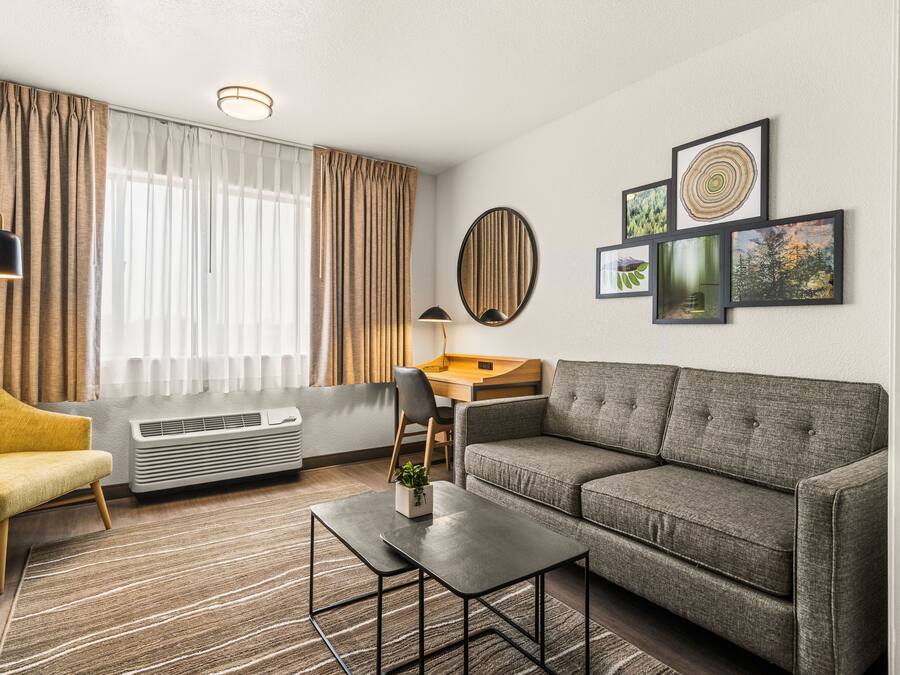 A modern hotel room with a grey sofa, a small table, a desk, a chair, and a large window with curtains.