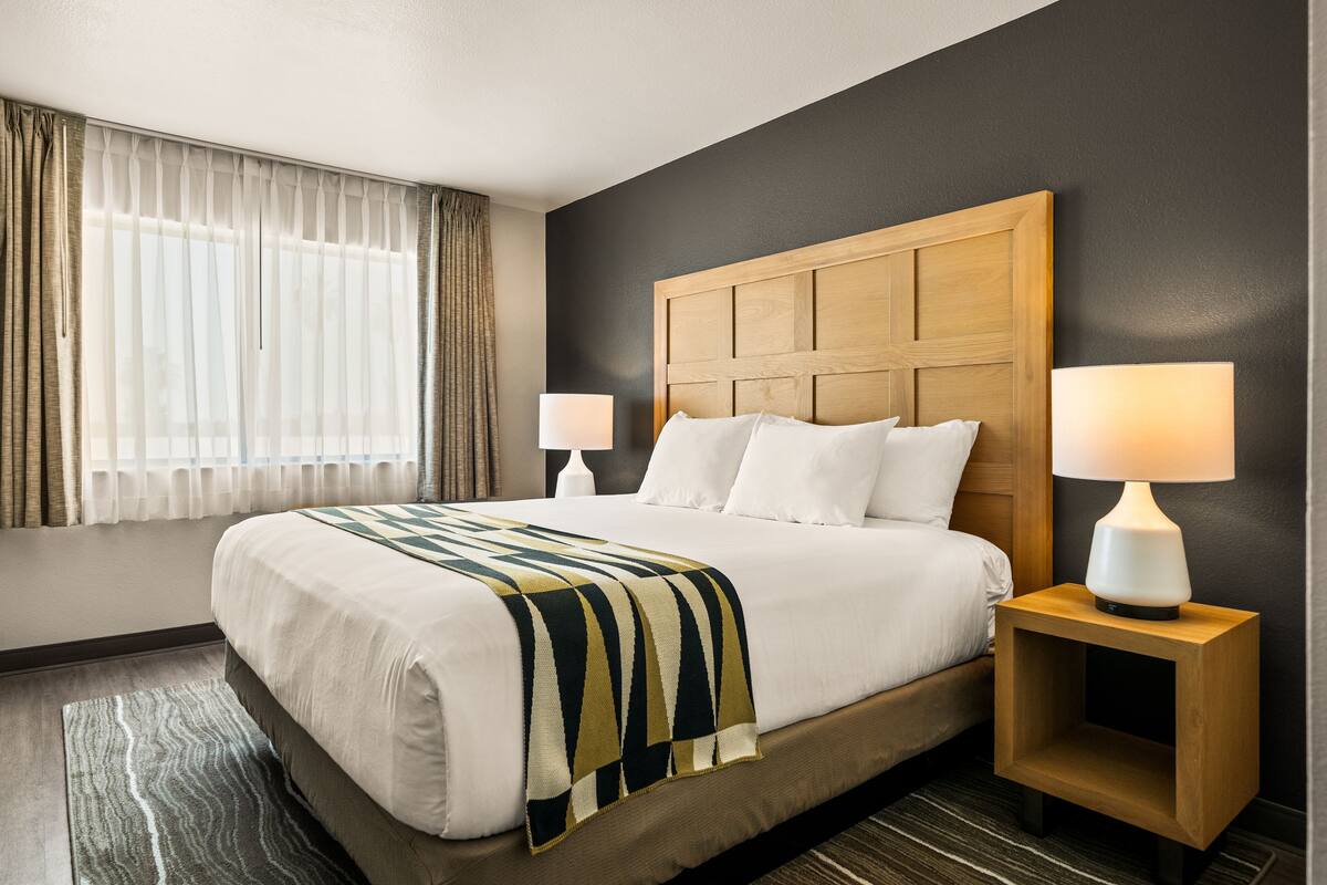 A hotel room with a large bed, two bedside tables with lamps, a window with curtains, and a dark wall.