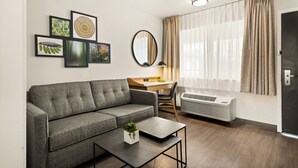 Un salon moderne avec un canapé gris, une petite table sur laquelle est posée une plante, un bureau avec une chaise, un grand miroir et une fenêtre avec des rideaux.