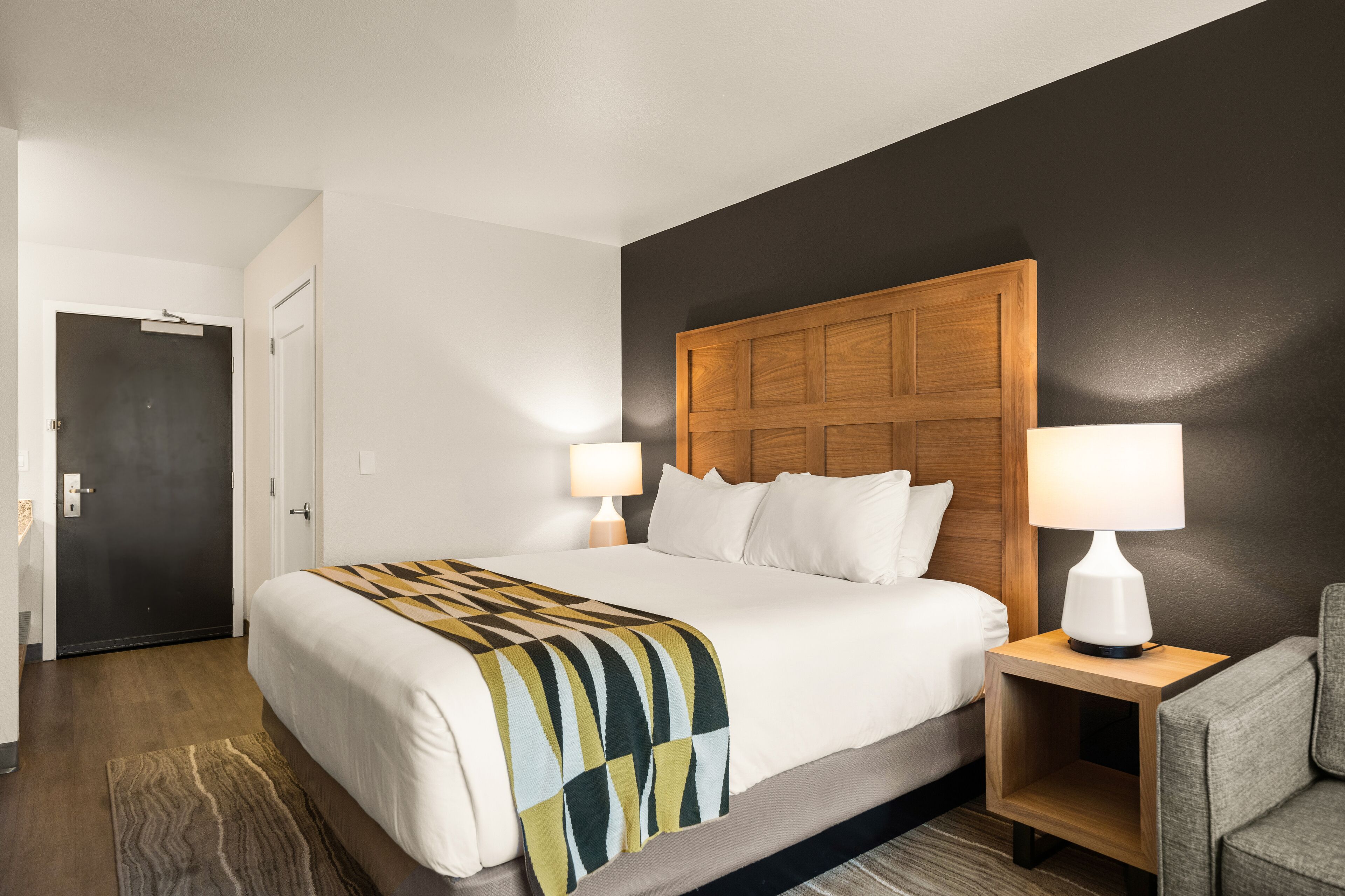 A hotel room with a large bed, two bedside tables with lamps, a wooden headboard, and a dark accent wall.