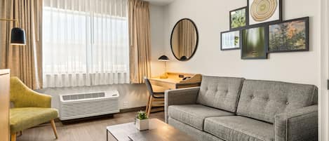 A modern living room with a grey sofa, a wooden desk, a chair, a small table with a plant, and a large window with curtains.