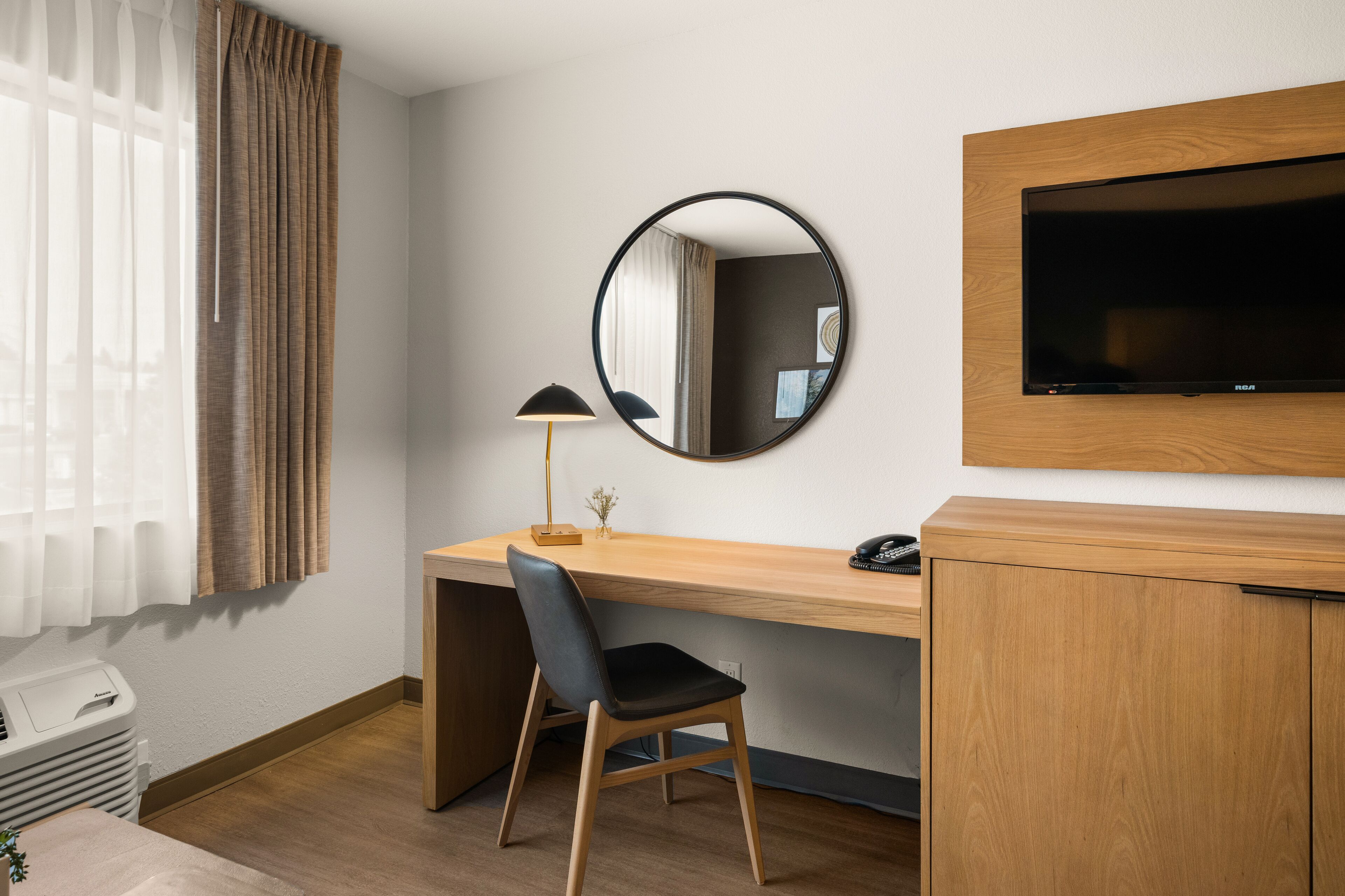 Habitación de hotel con un escritorio de madera, una silla, un espejo redondo, un televisor de pantalla plana y una ventana con cortinas.