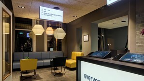 A hotel reception area with a modern design, including a reception desk, seating area with chairs, and a digital screen displaying room information. - a&o Rotterdam City (Rotterdam)