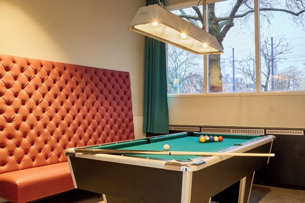 A pool table with balls, a red tufted bench, and a window with a view of trees and buildings.