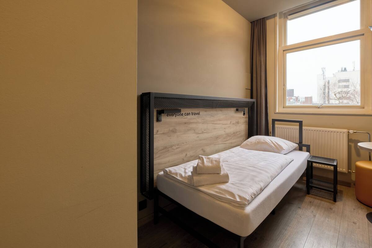 A single bed with a headboard, a small bedside table, and a window with a view of buildings.