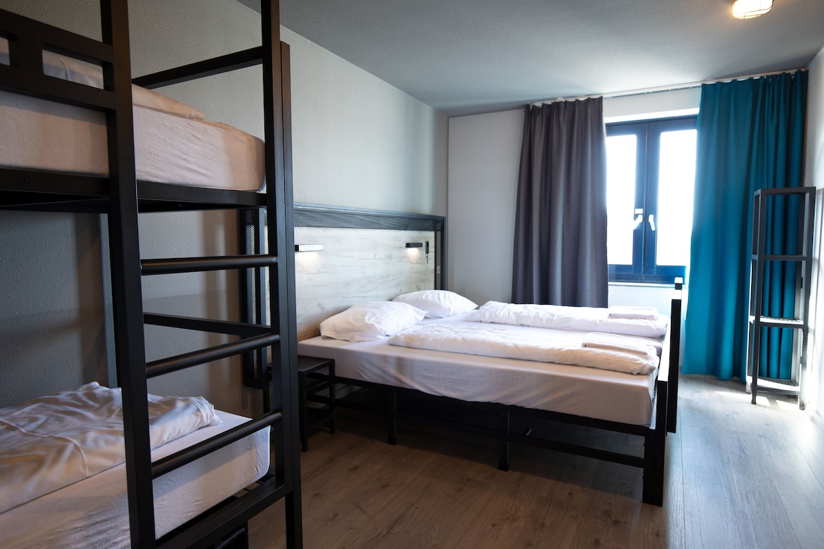 Ein Zimmer mit Etagenbetten, einem Einzelbett, einem Fenster mit Vorhängen und Holzboden.