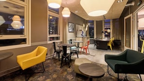 A modern hotel lobby with a mix of seating options, including yellow and green chairs, a small round table, and a bar area in the background. - a&o Rotterdam City (Rotterdam)
