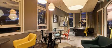 En moderne hotellobby med forskellige siddepladser, herunder gule og grønne stole, et lille rundt bord og et barområde i baggrunden.
