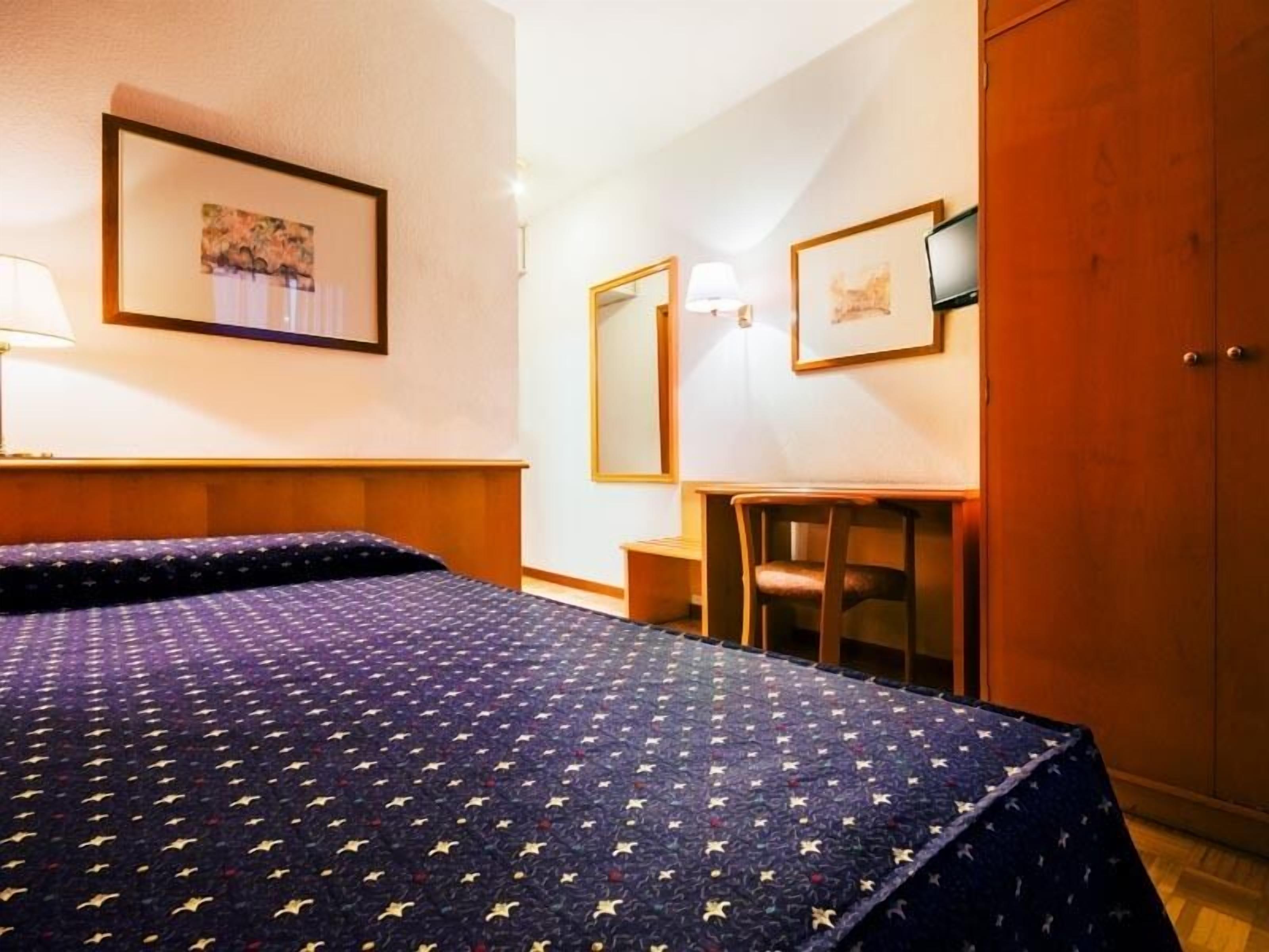 A hotel room with a bed, a desk, a chair, a wardrobe, and framed artwork on the walls.