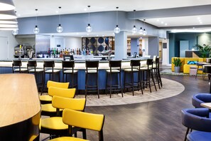 Un espace bar moderne doté d’un bar central, de nombreux tabourets et d’un coin salon avec des chaises jaunes.