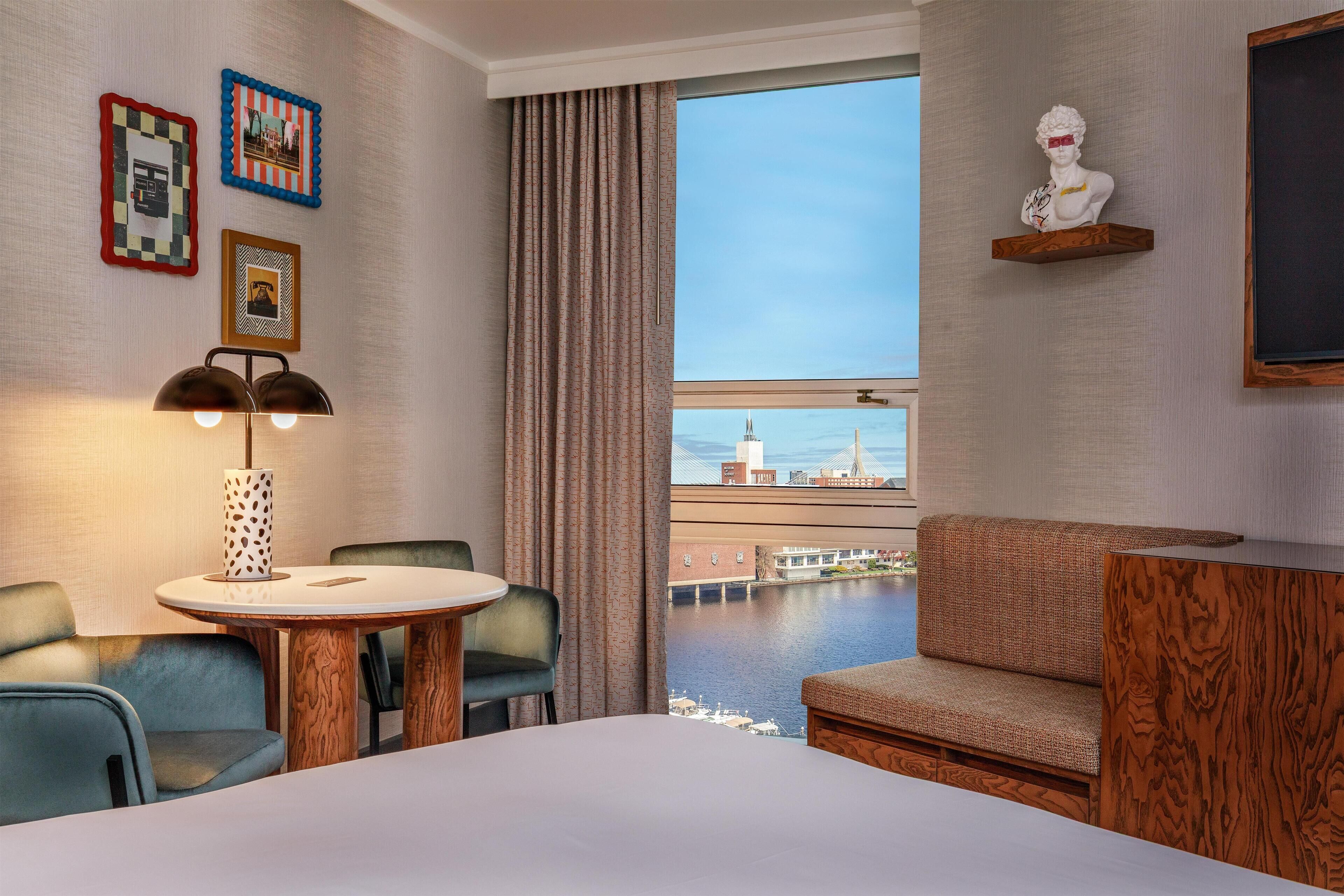 A hotel room with a round table, two chairs, a sofa, a large window with a city view, and a mounted bust on the wall.